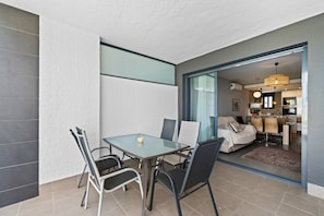 Ein modernes Wohnzimmer mit Glastisch und Stühlen, einer Couch und einer durch eine offene Tür einsehbarer Küche.
