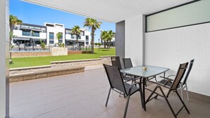 Une terrasse extérieure moderne, dotée d’une table et de chaises en verre, d’un mur en pierre et offrant une vue sur des immeubles et des palmiers.