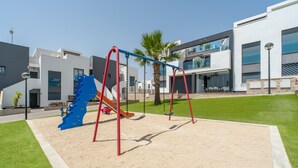 Une aire de jeux avec des balançoires et un toboggan, entourée d’une pelouse bien entretenue et de bâtiments modernes.