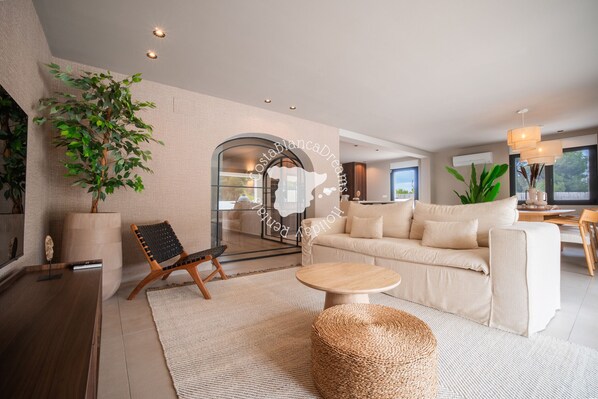A modern living room with a sofa, a coffee table, a chair, a large plant, and a glass partition. - CostaBlancaDreams - Villa Mariposa in Jávea (Javea)