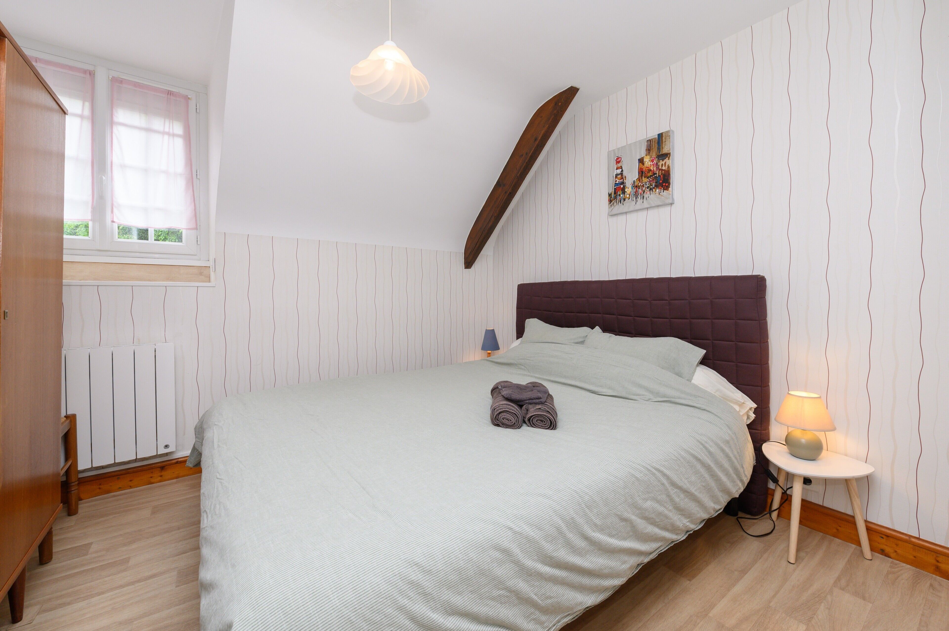 Une chambre à coucher avec un lit, une lampe de chevet, un radiateur, une fenêtre avec des rideaux et un tableau accroché au mur.