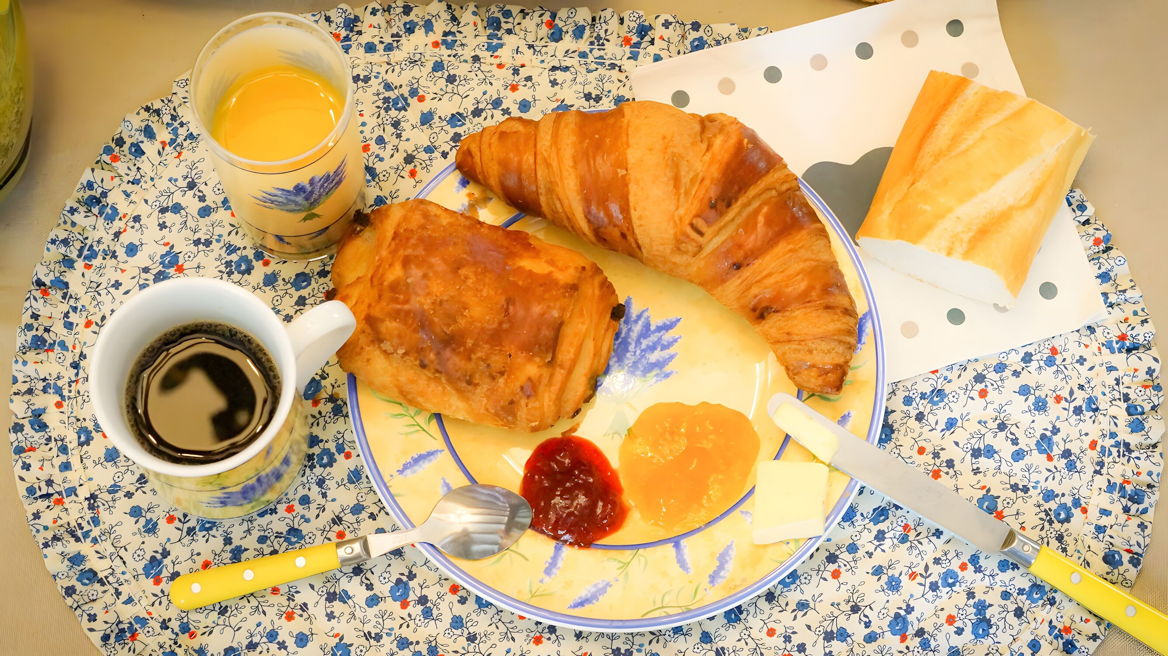 Ein Teller mit Croissants, Marmelade, Butter und Kaffee auf einer blumigen Tischdecke.
