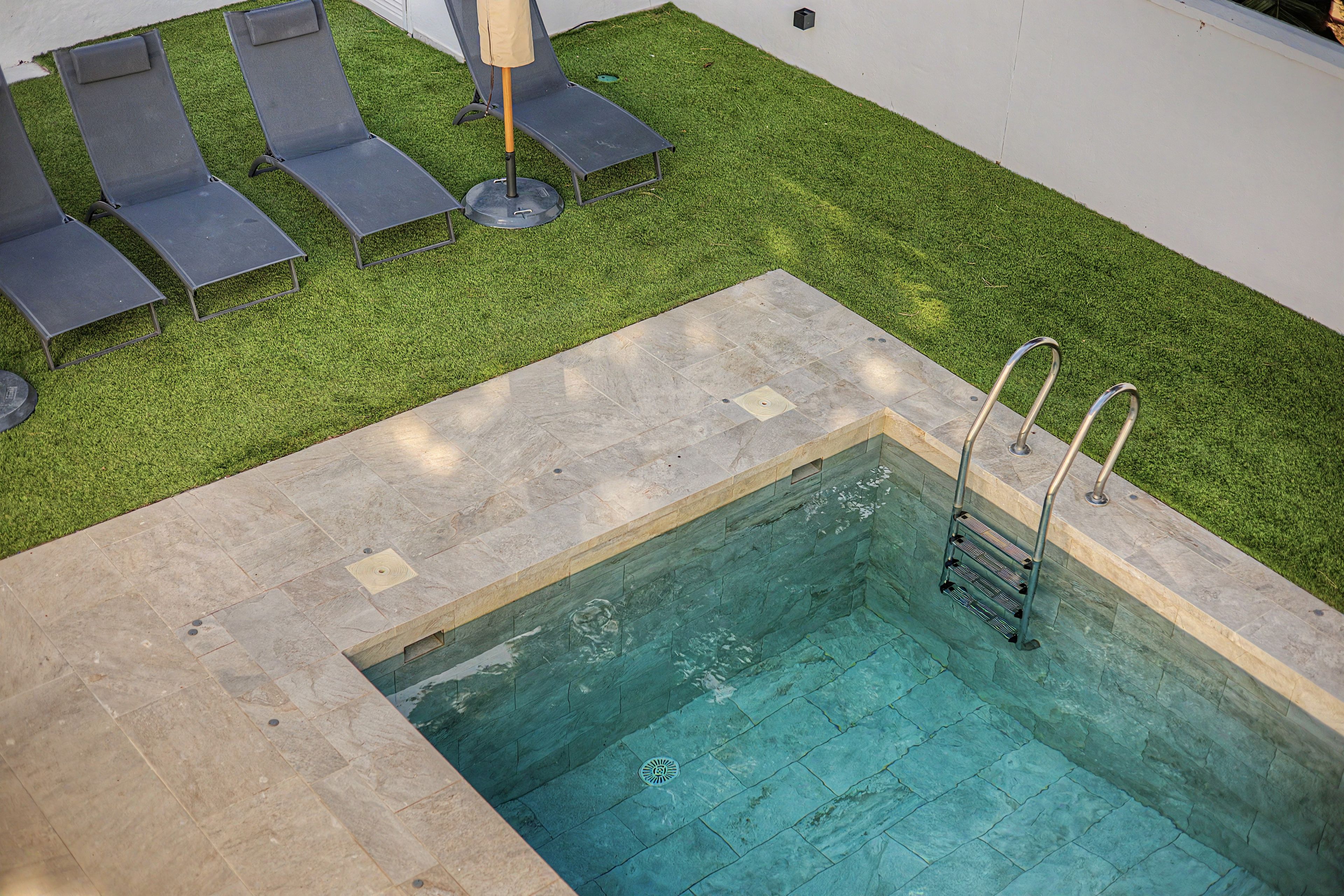 A swimming pool with a ladder, surrounded by a tiled deck and a grassy area with lounge chairs.