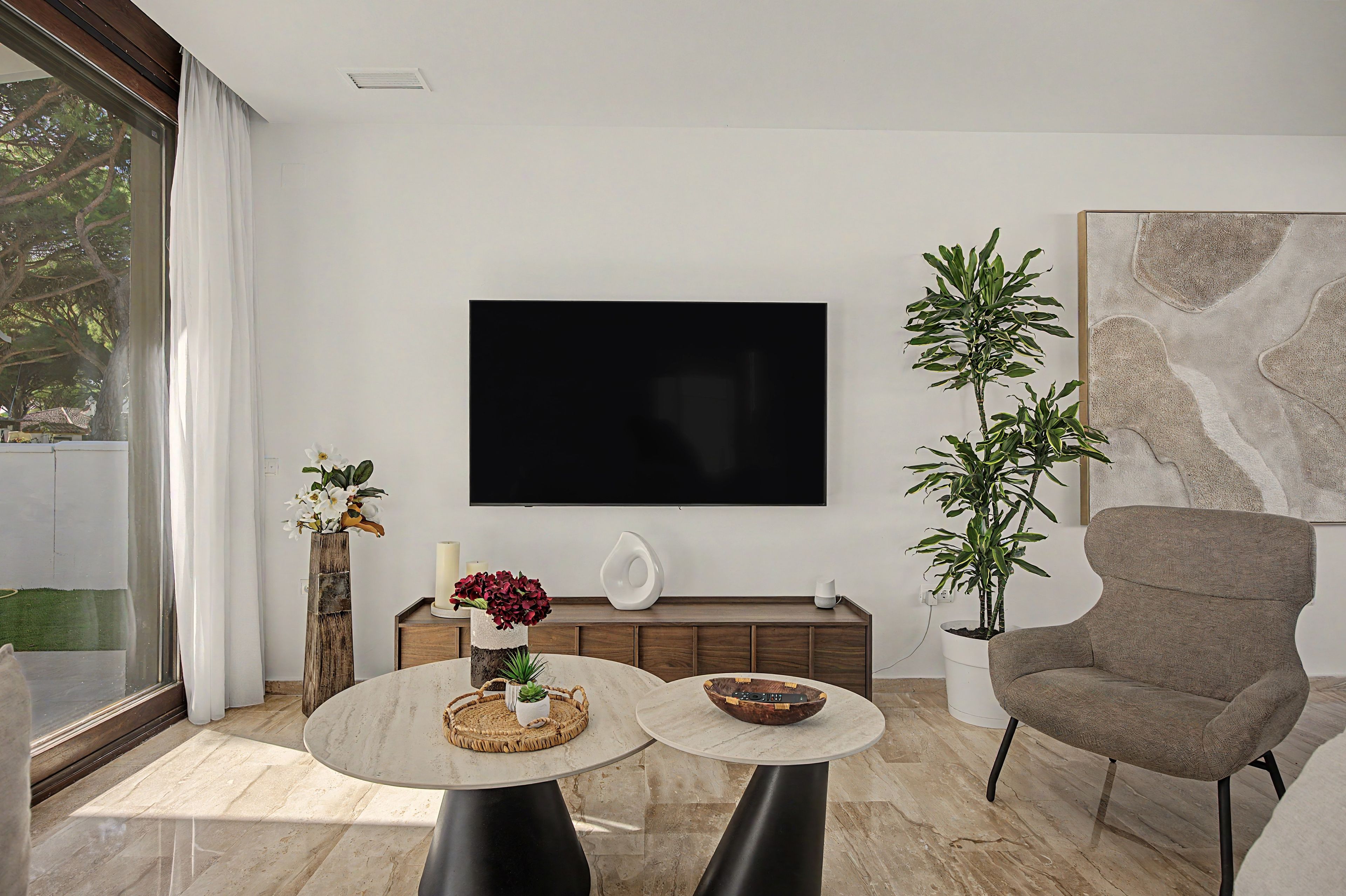 A modern living room with a large flat-screen TV mounted on the wall, a comfortable armchair, a round coffee table, and a potted plant.