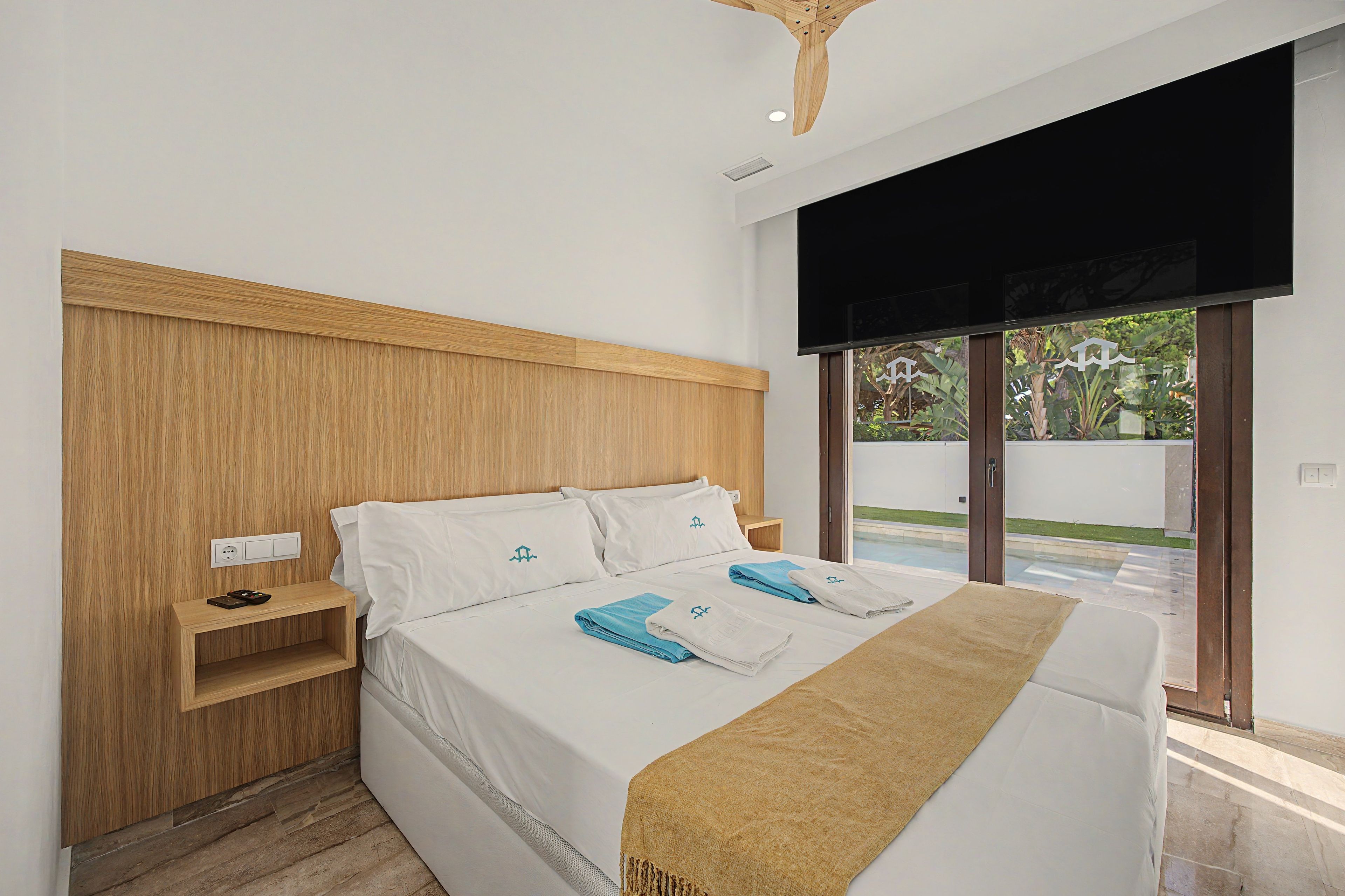 Una habitación de hotel moderna con cabecera de madera, una cama grande con ropa de cama blanca, vista al exterior a través de una puerta corrediza y un televisor de pantalla plana montado en la pared.