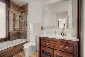 Un baño moderno con ducha envuelta en vidrio, inodoro blanco, tocador de madera con espejo y lavamanos.