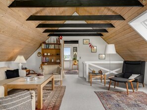 Une pièce de style « loft » avec un plafond en pente, des poutres en bois, un canapé, un fauteuil, une table, une bibliothèque et une vue sur l’extérieur grâce à une porte ouverte.