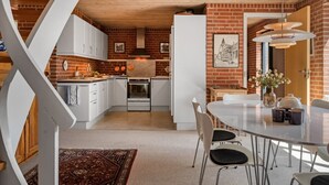 Une cuisine moderne avec des meubles blancs, un mur en briques, un coin repas doté d’une table en verre et un plafond en bois.