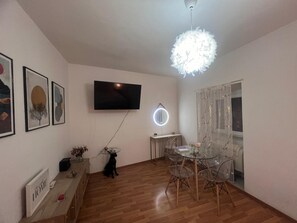 Ein Wohnzimmer mit Fernseher, einem runden Spiegel, einem Esstisch mit Stühlen und einer Deckenleuchte.