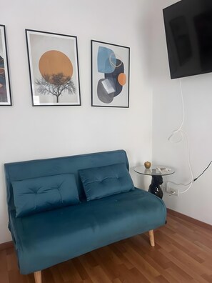 Ein türkisfarbenes Sofa mit zwei Kissen, ein Glastisch mit einem dekorativen Gegenstand und drei gerahmte Kunstwerke an der Wand.