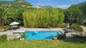 Una piscina con sillas de descanso y una zona de estar cubierta, rodeada de vegetación y montañas al fondo.