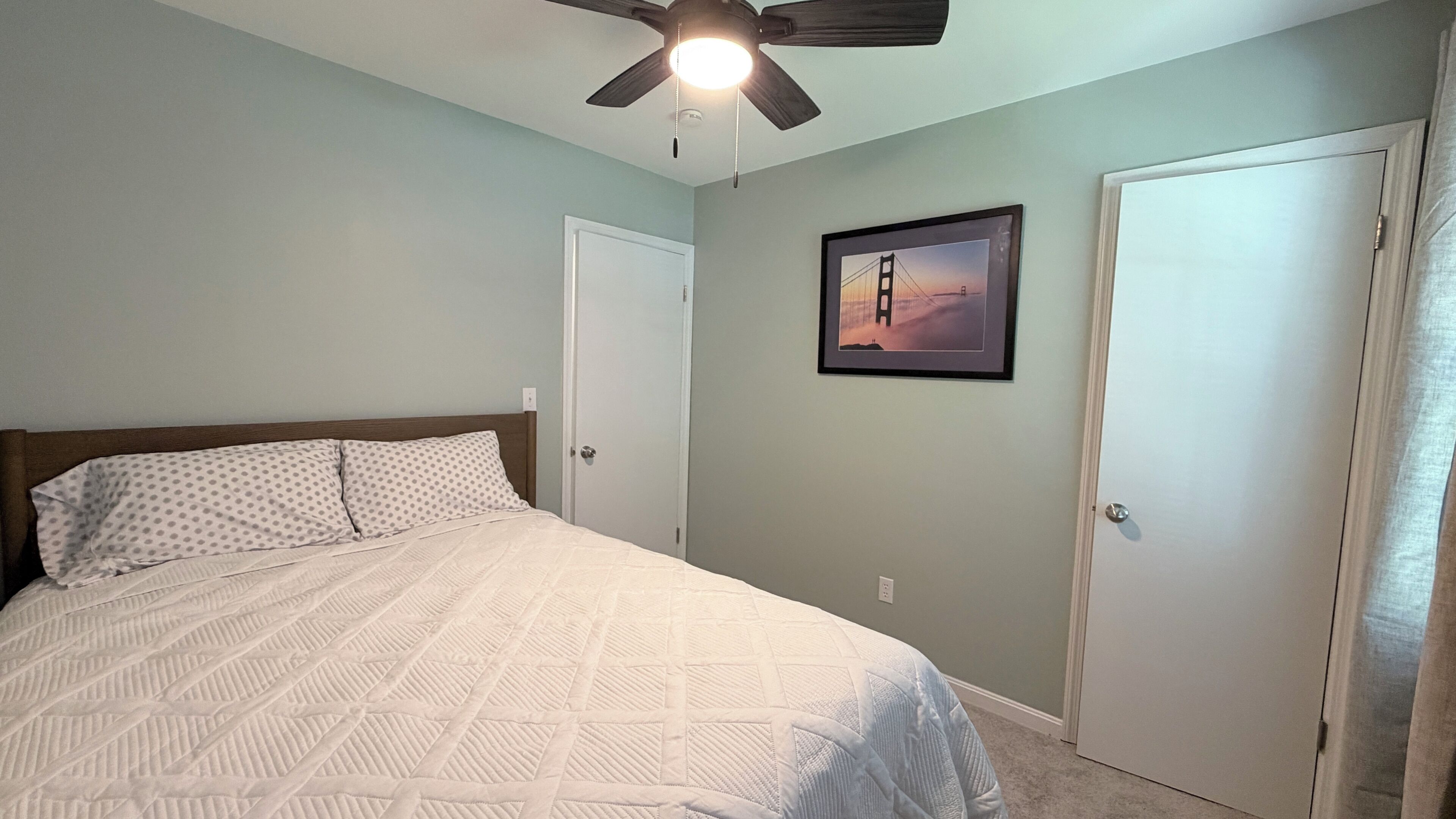 A bedroom with a bed, a ceiling fan, a door, and a framed picture on the wall.