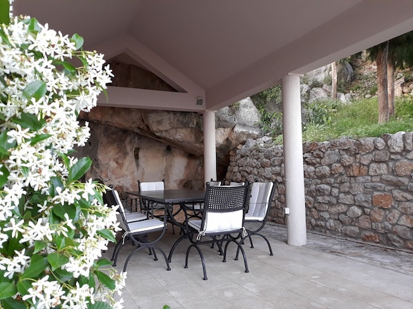 Eine überdachte Terrasse mit Steinmauer, Tisch und Stühlen sowie Blick ins Grüne.
