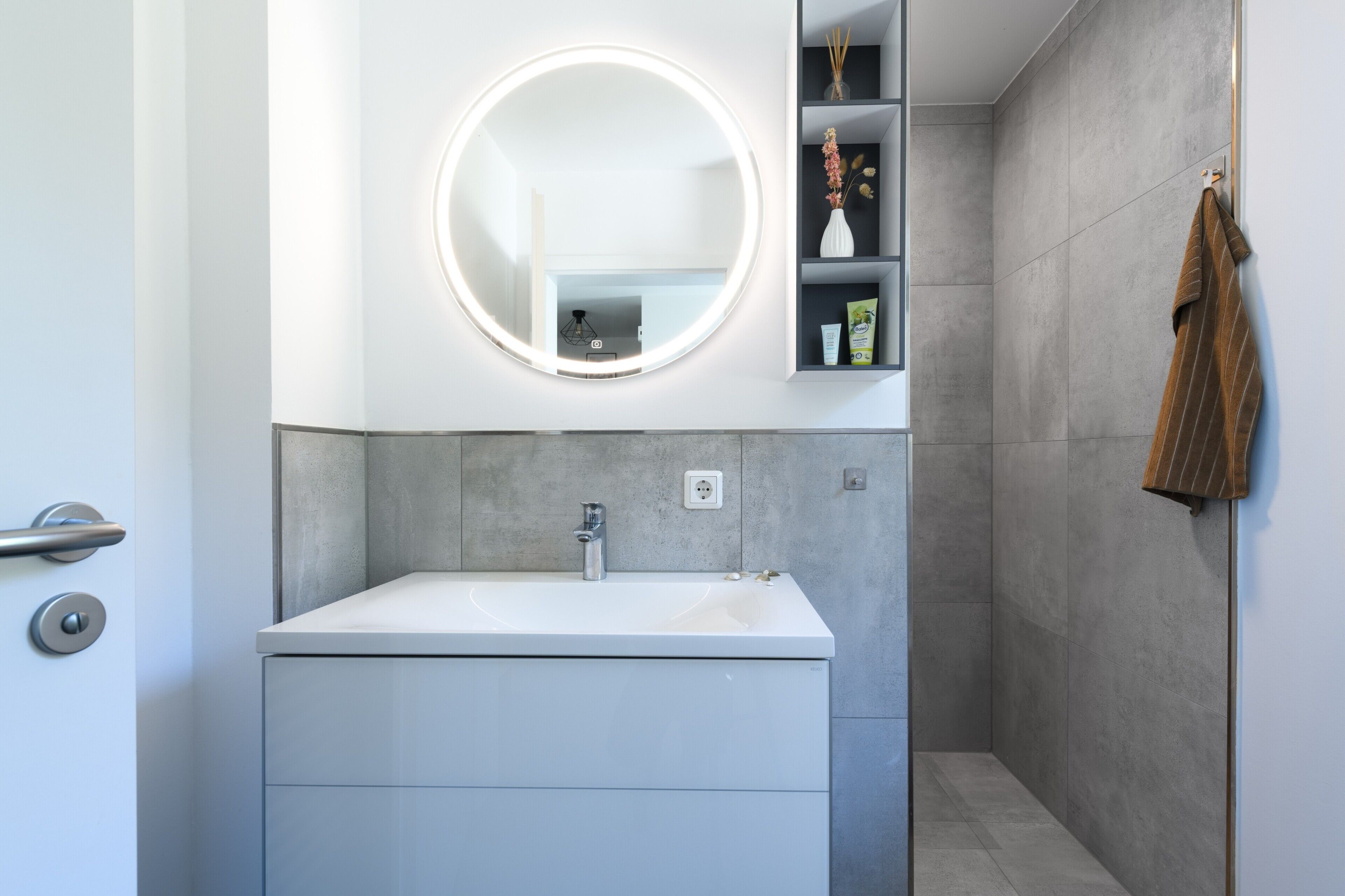 A modern bathroom with a white sink, a round mirror with lights, and a towel rack.
