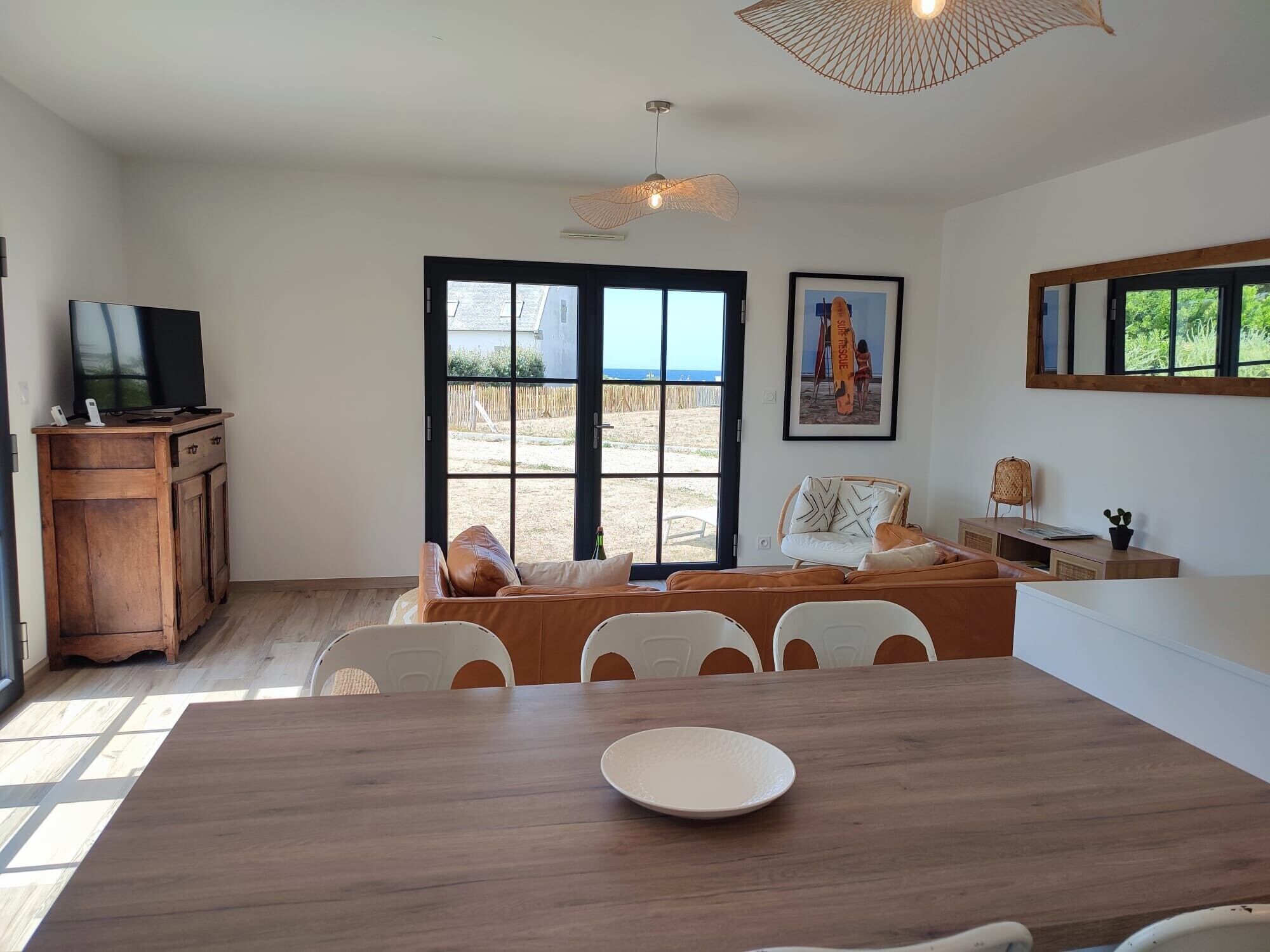 A modern living room with a wooden dining table, a television, and a beach view through large windows.