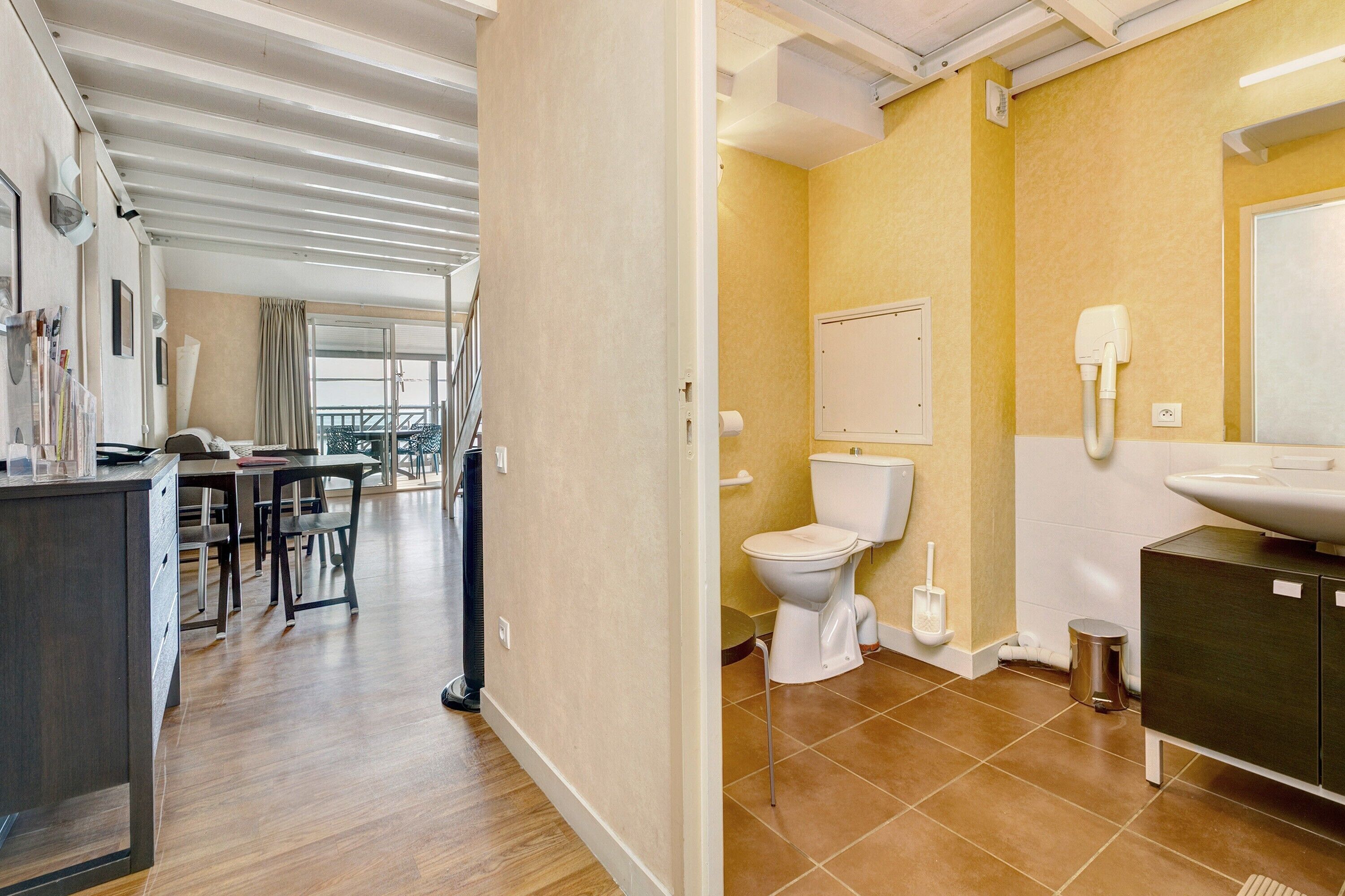 Quarto de hotel moderno com piso de madeira, área de jantar com mesa e cadeiras, banheiro com vaso sanitário e pia, e uma janela grande.