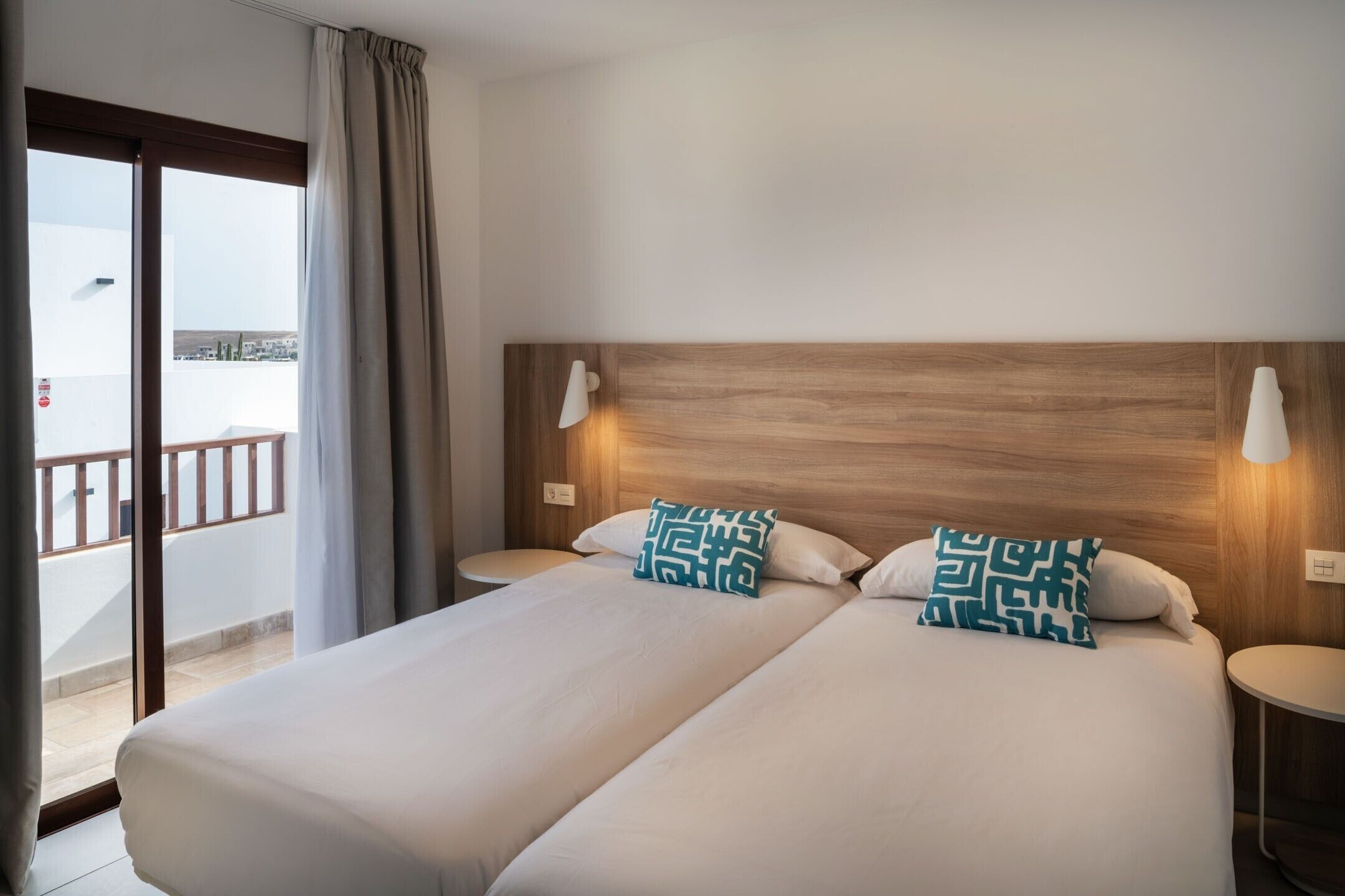 Une chambre d’hôtel avec deux lits, une tête de lit en bois, deux lampes de chevet, un balcon avec vue et une porte-fenêtre coulissante.