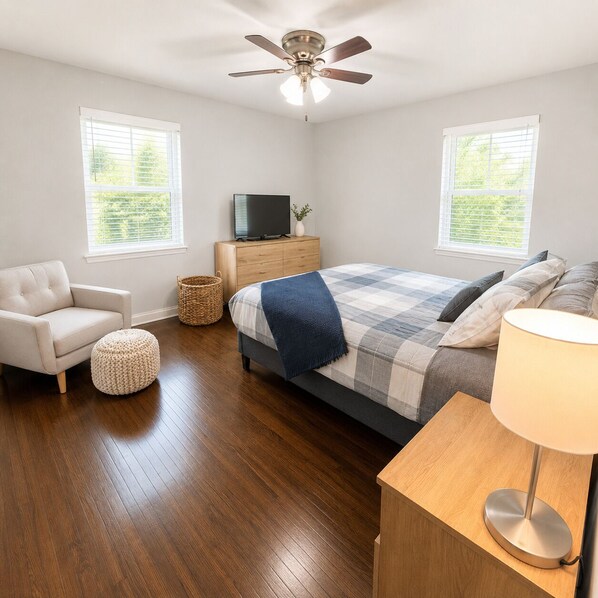 Une chambre à coucher comprenant un lit, une chaise, un téléviseur posé sur une commode en bois, un ventilateur de plafond et deux fenêtres.