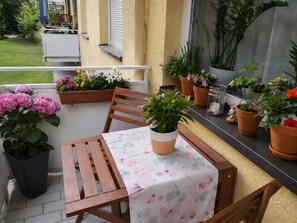 Ein Holztisch mit einer blumigen Tischdecke, umgeben von Topfpflanzen und einem Stuhl auf einem Balkon.