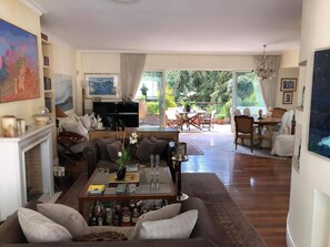A spacious living room with a fireplace, a dining area with a table and chairs, and a large window with a view of greenery. - Luxury Penthouse with Private Pool, Terrace, and Stunning Views (Athens)