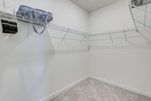 A corner of a room with a white wall, a grey carpet, and a metal wire shelf with a blue bag on it.