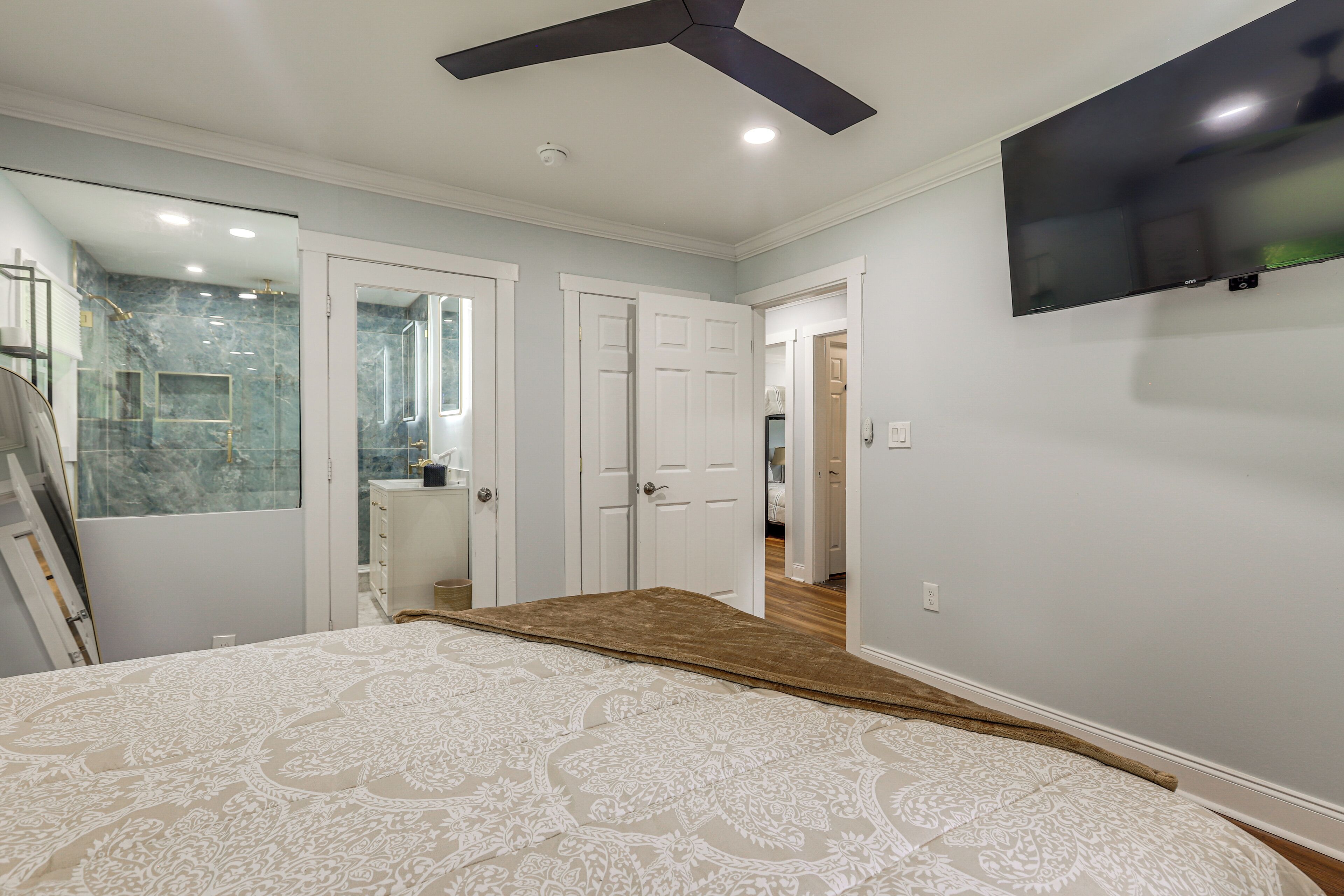 A bedroom with a large bed, a ceiling fan, a TV, and a bathroom with a glass shower.