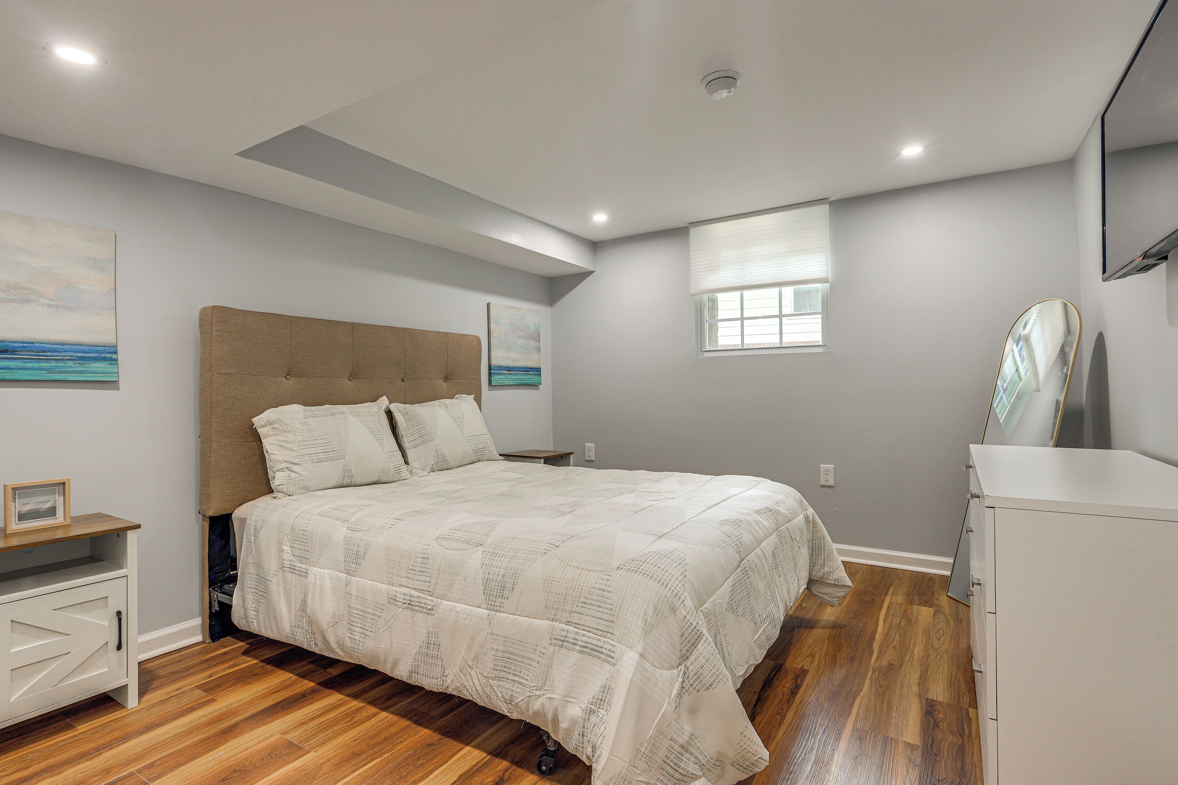 A bedroom with a bed, bedside tables, a dresser, a window, and artwork on the walls.