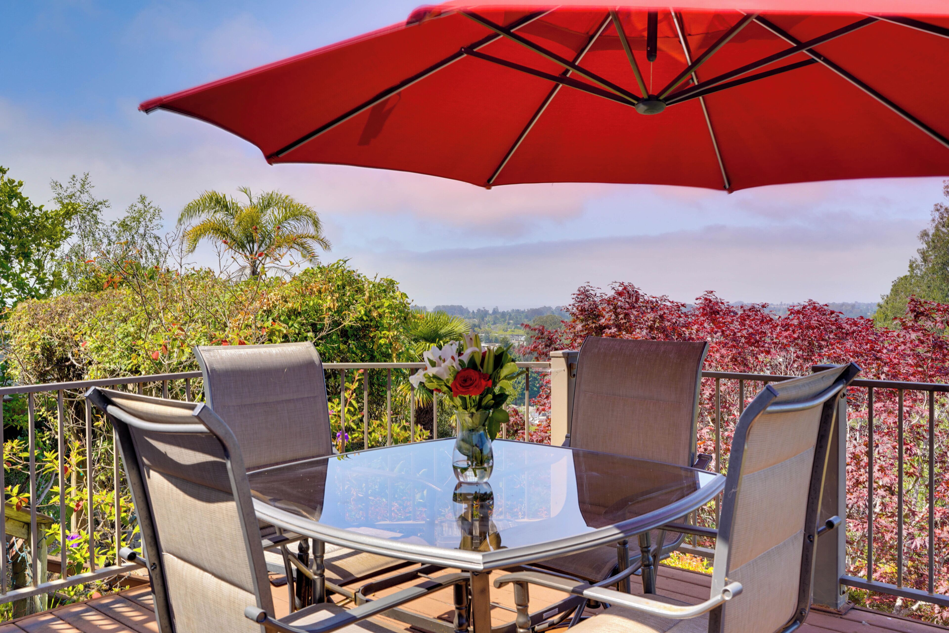 A round glass table with metal chairs and a red umbrella, set on a balcony with a view of trees and hills.