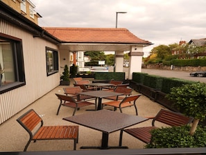 Eine Terrasse im Freien mit Tischen und Stühlen, ein überdachter Sitzbereich und ein Blick auf die Straße und die Gebäude im Hintergrund.