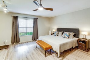 Un dormitorio con cama, banca, mesitas de noche, ventilador de techo y ventana con cortinas.