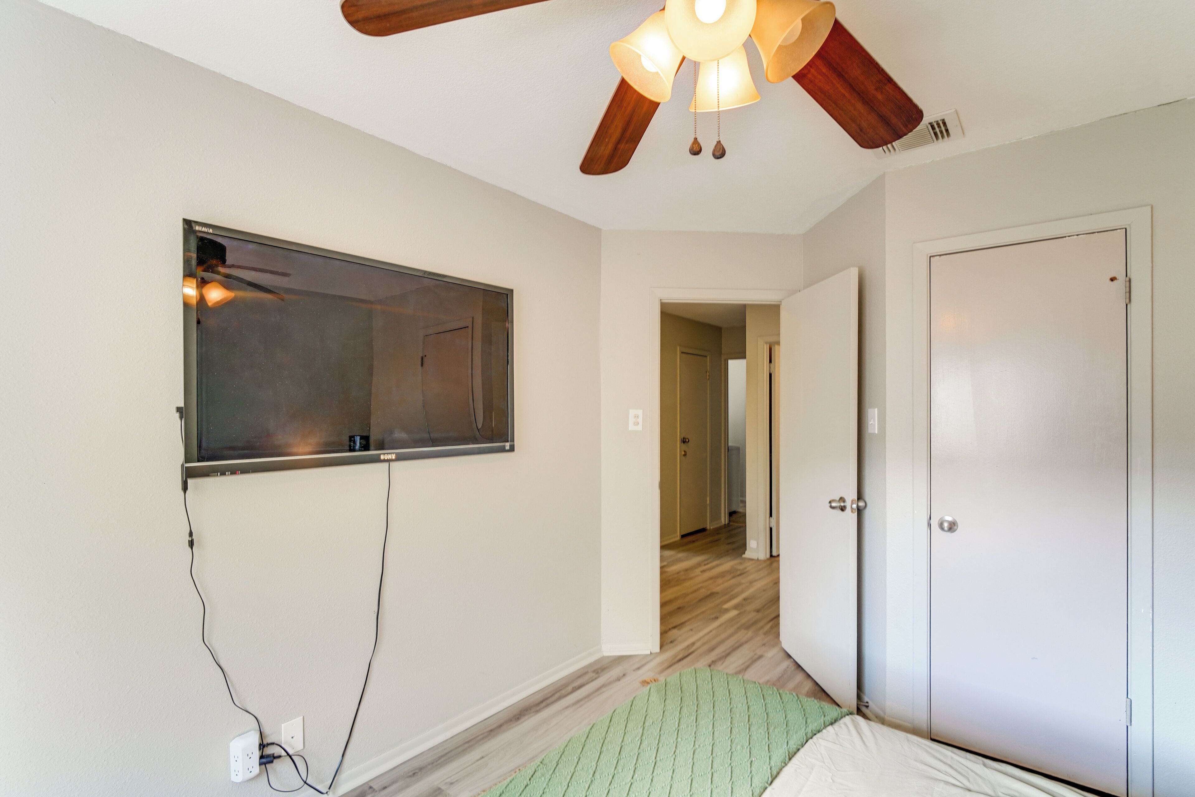 A room with a ceiling fan, a large window, a door, and a hallway leading to another room.