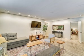 A modern living room with a sofa, a coffee table, a chair, a TV, a plant, and a small kitchen area.