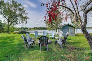 An outdoor seating area with Adirondack chairs and a table, a tree with red apples, and a small shed by a lake. - Historic Home on Beaver Dam Lake w/ Seasonal Dock (Cumberland)