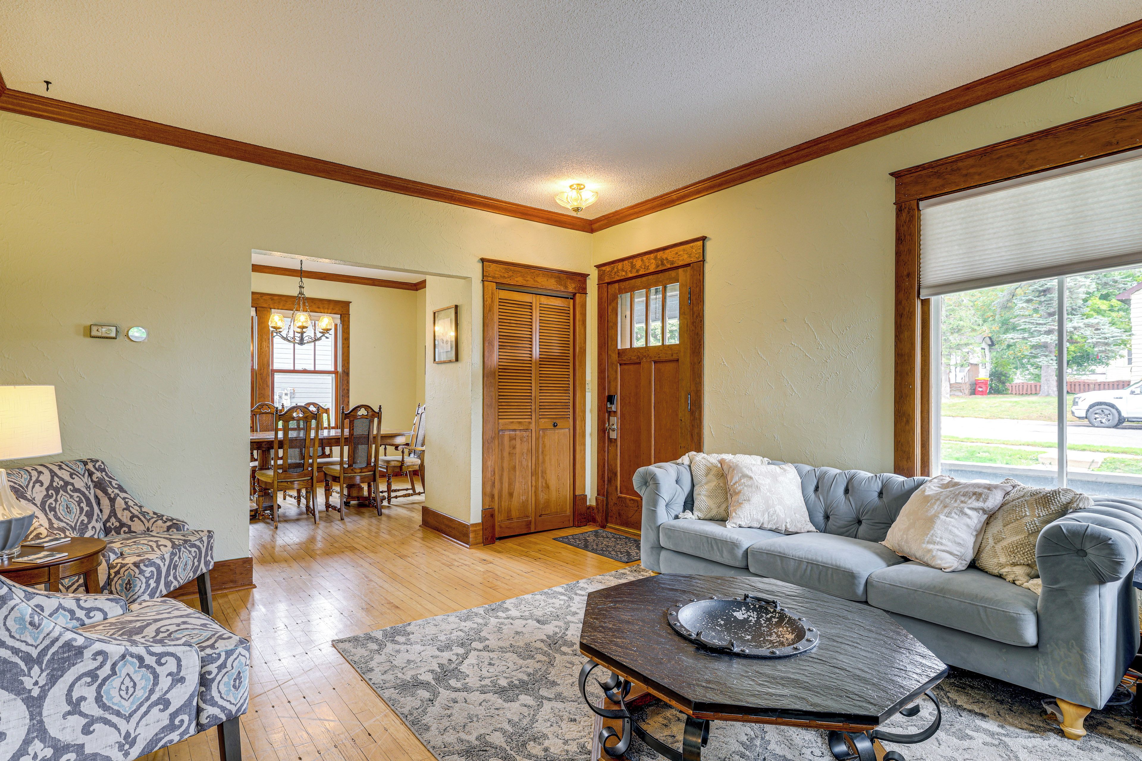 A living room with a grey sofa, patterned armchairs, a coffee table, and a dining area with wooden furniture.