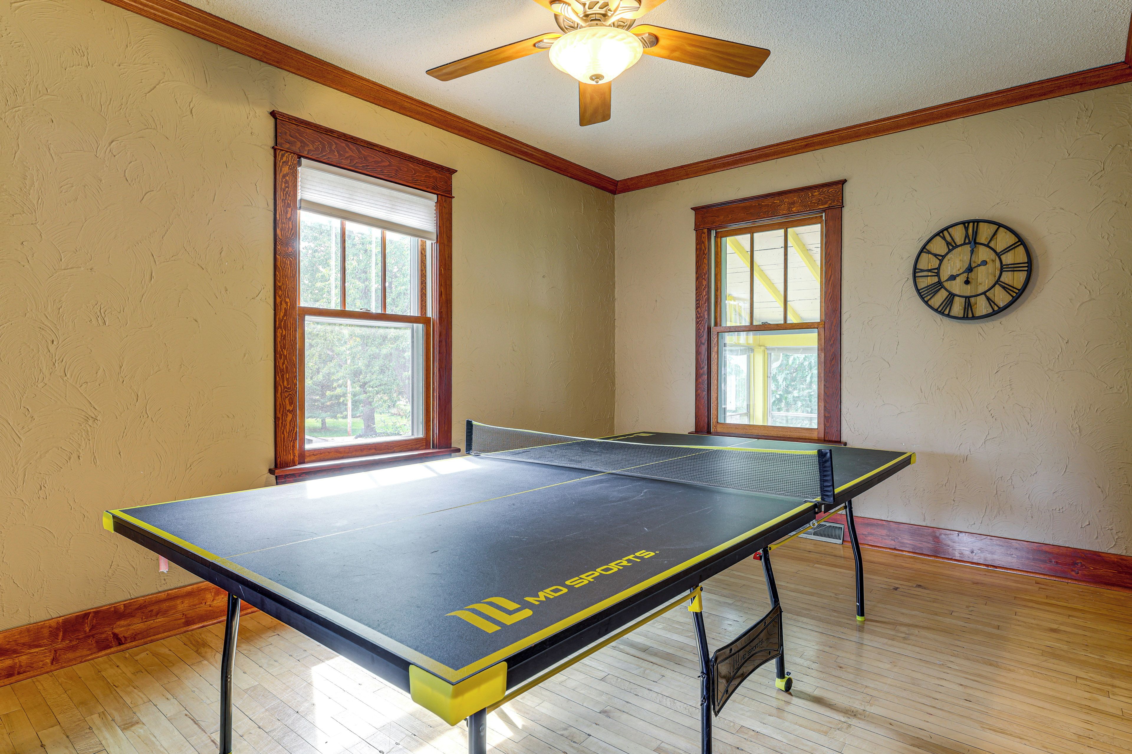 A room with a ping pong table, wooden flooring, two windows with blinds, a ceiling fan, and a wall clock.