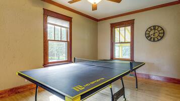 A room with a ping pong table, wooden flooring, two windows with blinds, a ceiling fan, and a wall clock.