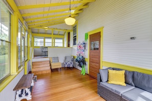 Quarto com piso de madeira, um sofá com travesseiro amarelo, uma cadeira, uma mesa e uma parede com porta.
