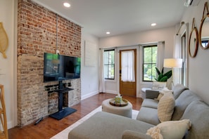 A living room with a brick wall, a flat-screen TV, a grey sofa, a coffee table, and a door with a sign. - 2 Mi to Dtwn Knoxville! Dog-friendly Apt (Knoxville)