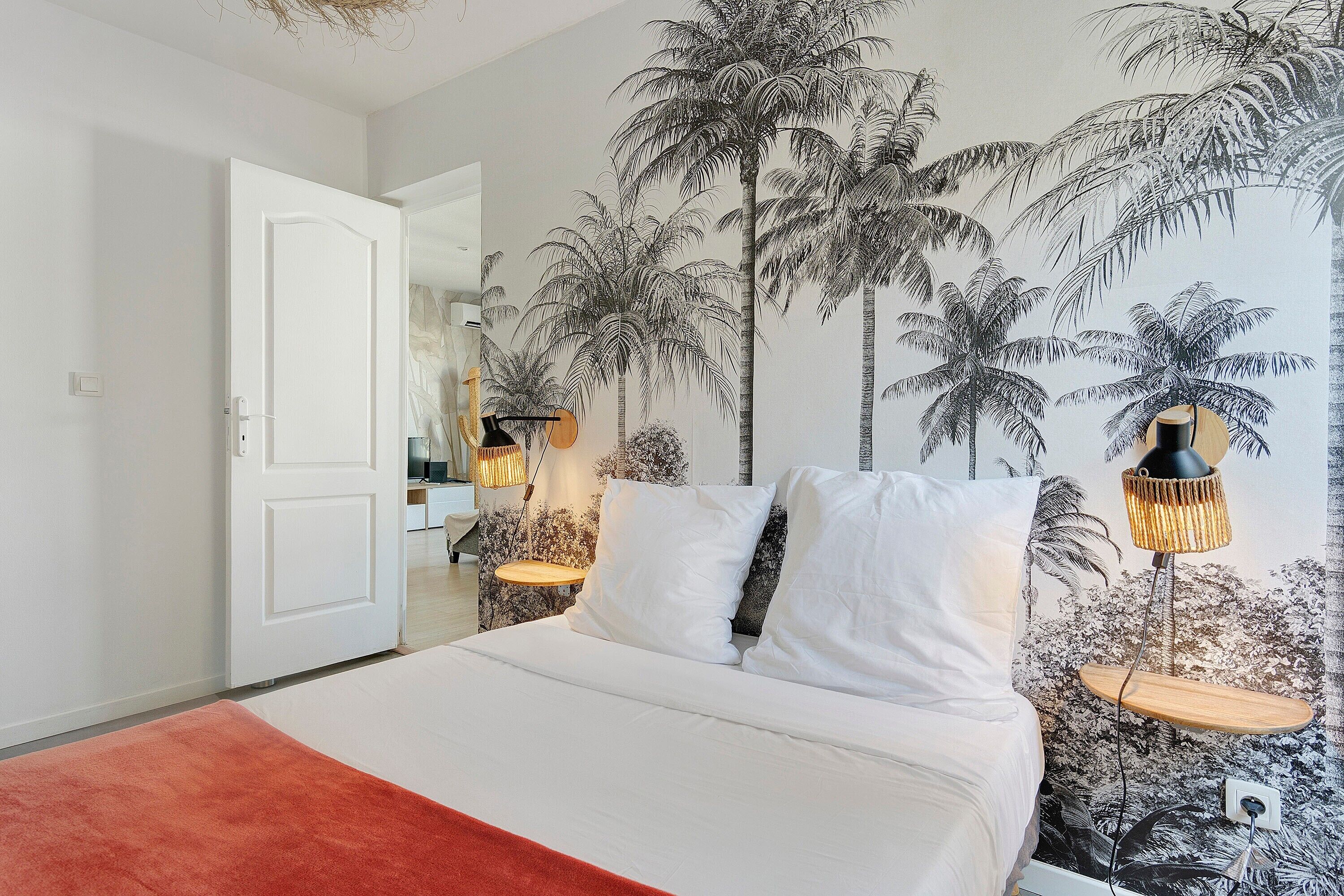Ein Schlafzimmer mit einem Bett, weißen Kissen, einer roten Decke, einer Wandmalerei von Palmen und zwei Nachttischlampen.
