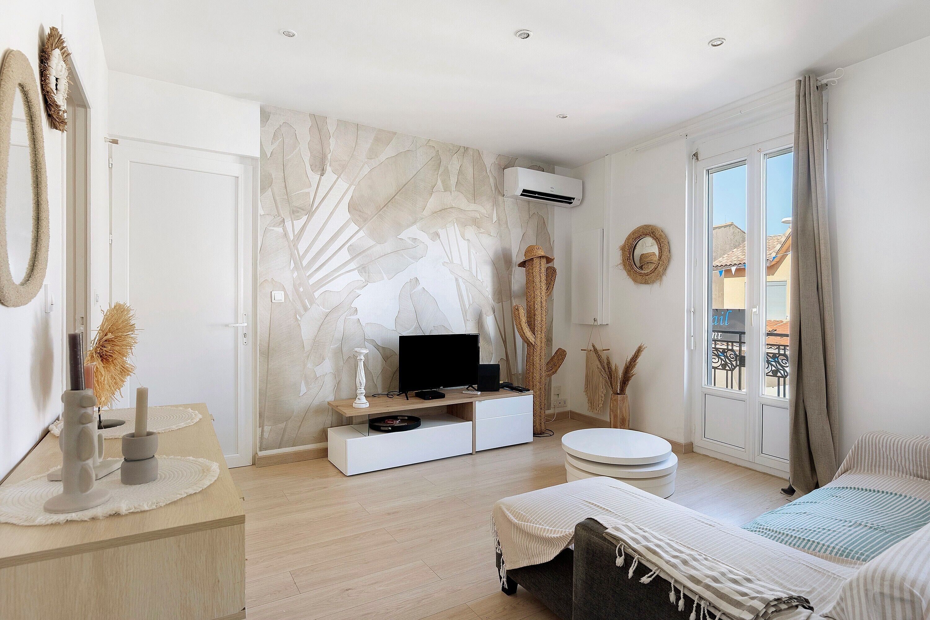 A modern bedroom with a flat-screen TV, a white sofa, a round ottoman, a cactus, and a balcony with a view.
