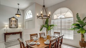 Un comedor con una mesa de madera dispuesta para seis personas, rodeada de sillas, con una araña colgante y un espejo grande en la pared.