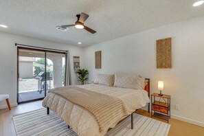 Quarto com cama, ventilador de teto, mesa de cabeceira com abajur, porta de vidro deslizante e uma planta.