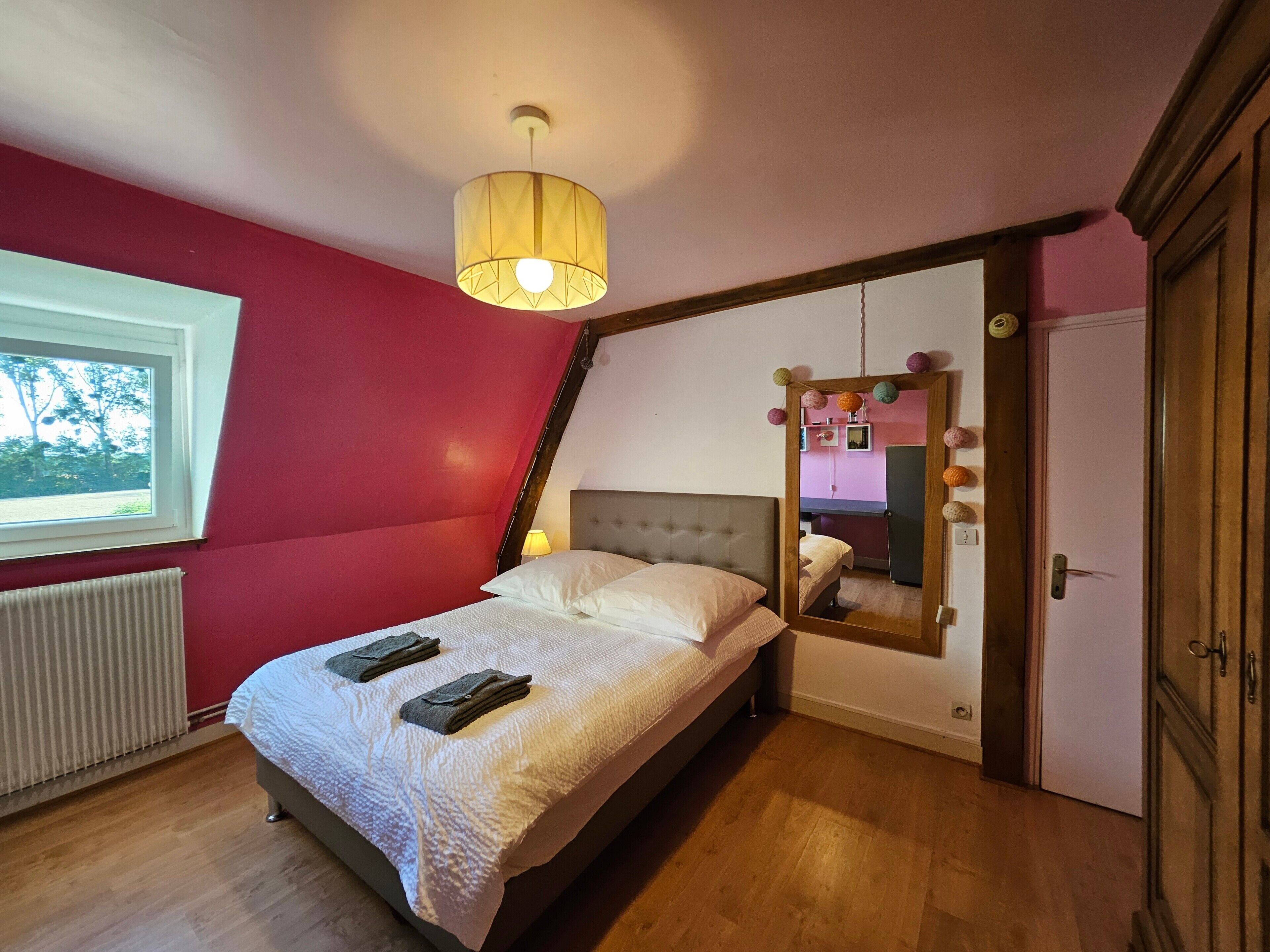 A bedroom with a bed, a window, a radiator, a mirror, and a door.