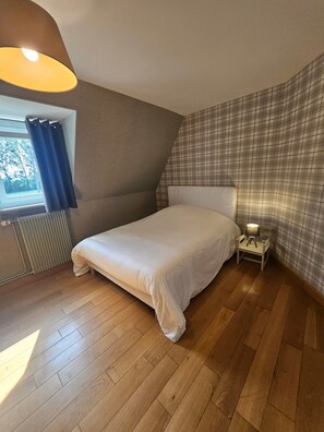 Et soveværelse med en seng, et natbord, en lampe, et vindue med gardiner, en radiator og en væg med tern.