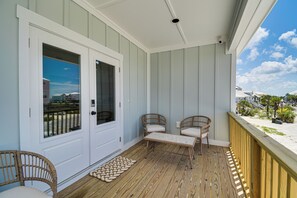 Ein überdachter Balkon mit einer weißen Tür, zwei Korbstühle mit Kissen, einem kleinen Tisch und einem Holzboden.