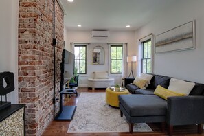A living room with a brick wall, a gray sofa, a yellow ottoman, a white armchair, and a large window. - Extended East Knoxville Stay, 2 Mi to Dtwn! (Knoxville)