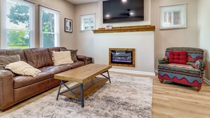 Una sala de estar con un sofá de cuero marrón, una mesa de centro de madera, un sillón colorido, una chimenea y un televisor de pantalla plana montado en la pared.