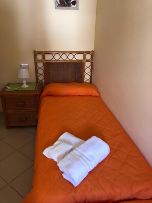 Uma cama com colcha laranja e toalha branca, mesa de cabeceira com abajur e cabeceira de vime.