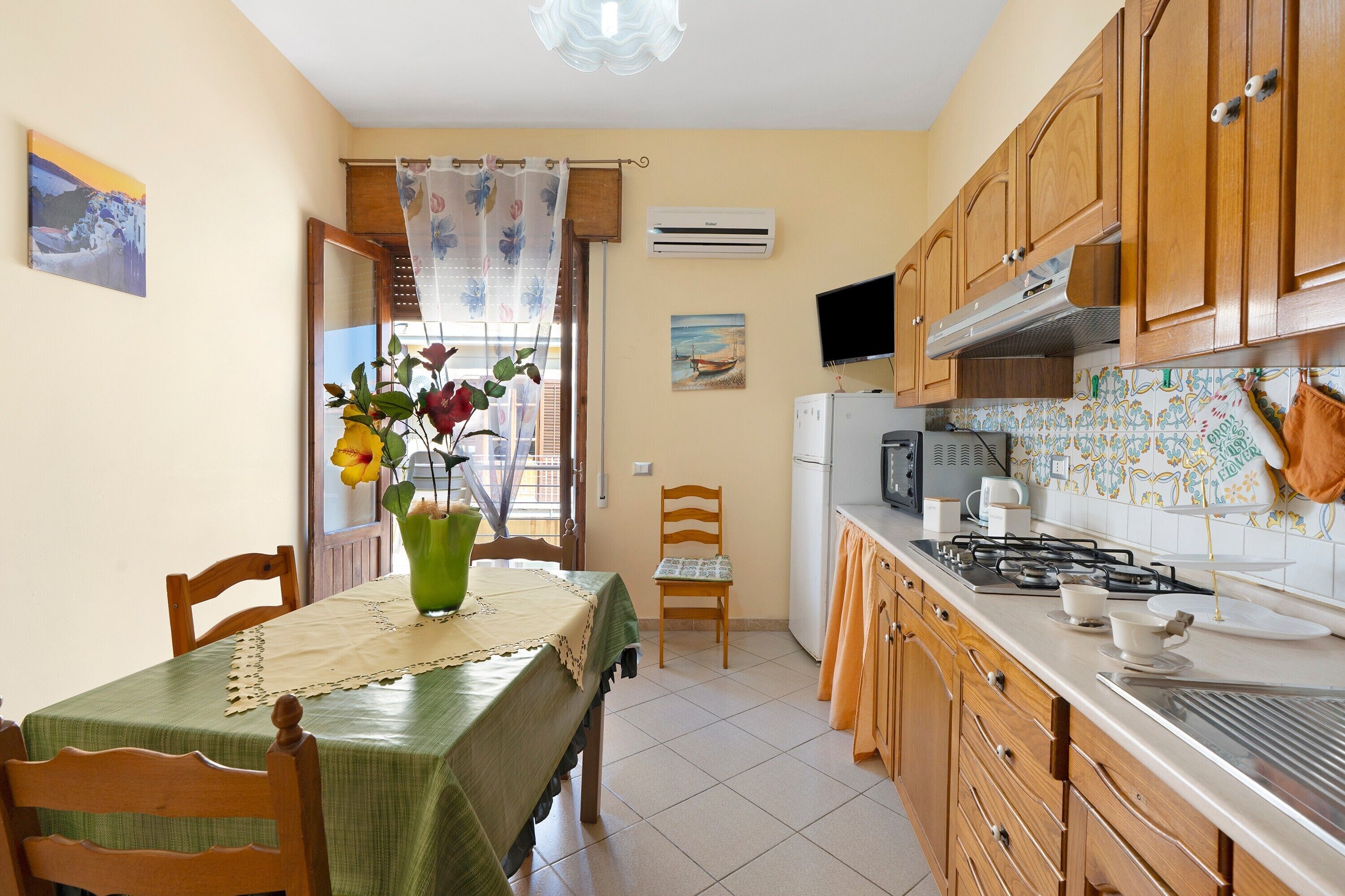 A kitchen with wooden cabinets, a dining table with a green tablecloth, a chair, a vase with flowers, and a window with a curtain.