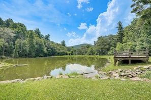 A serene lakeside setting with a wooden bridge, lush greenery, and a clear sky.